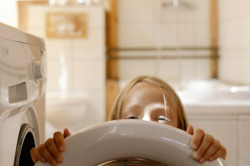 Barn som tittar fram bakom tvättmaskinen. Hur mycket tvättmedel ska man använda och hur mycket sköljmedel ska man använda